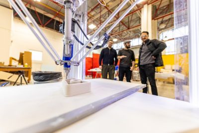 A close-up view of a 3D printer in action, extruding layers of material to form a rectangular object, with three researchers standing in the background observing the process in a brightly lit workshop.