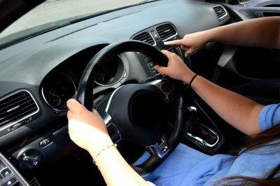 Two people sitting inside of a car, one has their hands on the steering wheel while the other is pointing at the dashboard